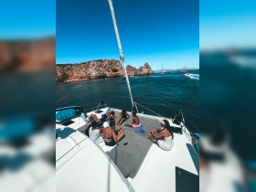 A lively group enjoys sunbathing and relaxation on the deck of a catamaran, surrounded by stunning coastal scenery — perfect for a fun day out on the water.