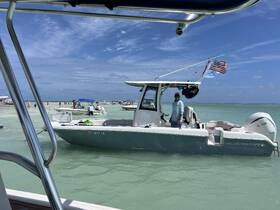 A man stands on the deck of a Sea Hunt boat in shallow waters, surrounded by other boats and beachgoers enjoying a sunny day on the water.