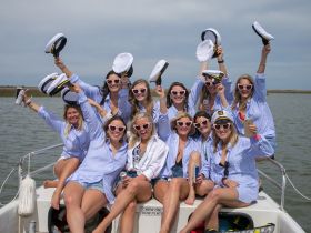 A lively group of friends celebrates on a boat, wearing captain hats and heart-shaped sunglasses, perfect for a fun day on the water.
