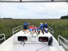 A group of children in life jackets lean over the bow of a boat, eagerly exploring the lush marshland ahead — a perfect moment of adventure and curiosity on the water.
