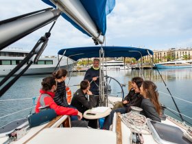 A group of guests enjoys a sailing lesson on a yacht, engaging with the instructor while surrounded by beautiful marina views.