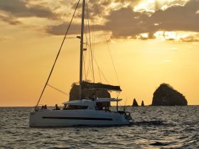 A stunning catamaran sails into the sunset, showcasing its elegant design against a picturesque backdrop of rocky islands — perfect for sunset cruises and sailing adventures.