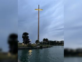 A view of a large illuminated cross near the water, surrounded by palm trees and a cloudy sky. The scene captures a serene atmosphere, but details are not clearly defined.