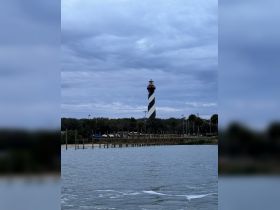 A distant view of a lighthouse against a cloudy sky, with a dock and boats in the foreground. The image lacks clarity and detail.