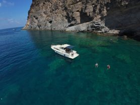 A scenic view of a boat anchored near a rocky coastline, with people enjoying a swim in the crystal-clear waters — a perfect spot for summer adventures.