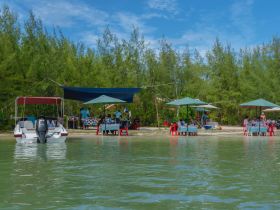 A lively beach scene featuring a group enjoying a meal under umbrellas, with a boat anchored nearby — perfect for outdoor dining experiences by the water.