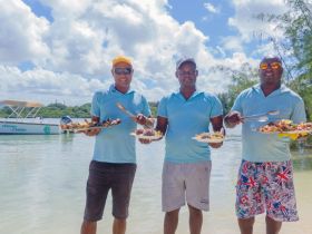 Three friendly staff members serve delicious local dishes at a scenic beach location, showcasing the culinary delights of the cruise experience.