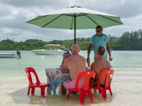 A group enjoys drinks at a beachside table under an umbrella, with boats anchored nearby, capturing the essence of a relaxing day on the water.