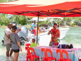 A lively group enjoys a beachside gathering with a boat in the background, capturing moments of fun and celebration under a vibrant tent.