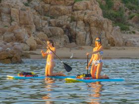 Two women enjoy paddleboarding in serene waters near a beach, capturing the essence of summer fun and adventure on the water.