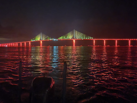 A nighttime view of a brightly lit bridge reflecting on the water, creating a stunning visual effect. The image captures the ambiance of a serene evening on the water.
