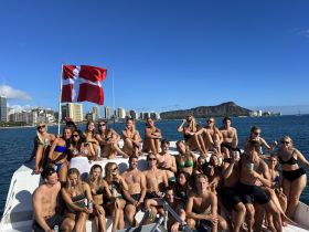 A vibrant group of friends enjoys a fun day on the water, celebrating with drinks aboard a boat, with stunning views of the coastline and mountains in the background.