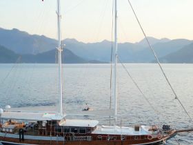 A stunning aerial view of a yacht docked in serene waters, with a person in a small boat nearby, set against a backdrop of majestic mountains.