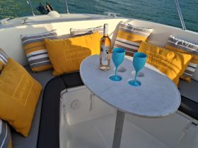 A stylish setup featuring colorful cushions, a bottle of wine, and blue glasses on a boat table, perfect for relaxing and enjoying drinks on the water.