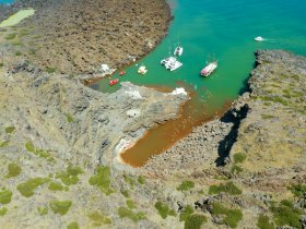 A stunning aerial view of a secluded cove with multiple boats anchored amidst rocky terrain and vibrant turquoise waters, showcasing the beauty of coastal adventures.
