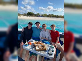 A joyful family enjoys a meal on a boat, surrounded by stunning turquoise waters and a sandy beach backdrop — perfect for memorable family outings.