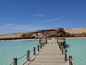 A scenic view of a wooden pier leading to a beachside establishment, surrounded by clear turquoise waters and a mountainous backdrop.