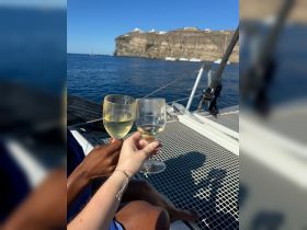 A couple toasts with glasses of wine aboard a yacht, enjoying a beautiful day on the water with stunning coastal scenery in the background.