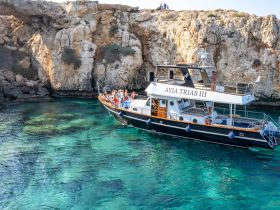 A lively group enjoys a scenic boat trip aboard the AYIA TRIAS III, surrounded by stunning rocky coastlines and crystal-clear waters.