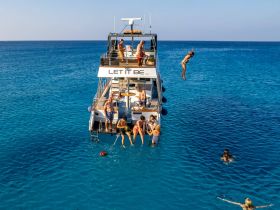 A lively group enjoys a day on the water, with one person diving off the boat into the crystal-clear sea — a perfect scene for summer fun and adventure.