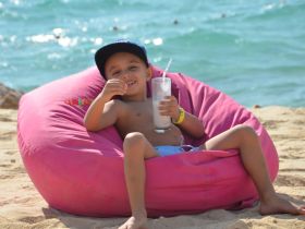 A cheerful child enjoys a refreshing drink while lounging on a pink bean bag at the beach, capturing the essence of relaxation and fun in the sun.