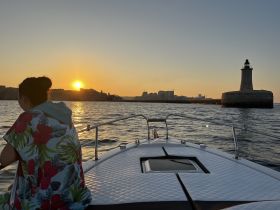 A serene sunset view from a boat, with a silhouette of a person enjoying the scenery and a lighthouse in the background — perfect for romantic evening cruises.