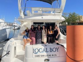 A lively group of friends poses on the deck of the 'Local Boy' yacht in Cabo San Lucas, celebrating with drinks and enjoying their time on the water.