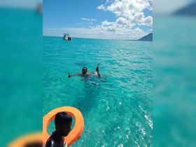 A lively scene in the turquoise waters, featuring a man enjoying drinks while swimming, with a child nearby and a boat in the background — perfect for a fun day on the water.