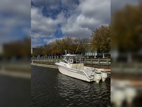 A Grady-White boat docked along a serene waterfront, showcasing its sleek design against a backdrop of autumn foliage and cloudy skies.