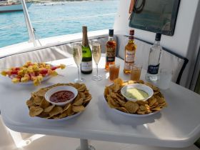 A vibrant spread of snacks and drinks on a boat table, featuring fruit skewers, chips with salsa, and a selection of spirits — perfect for a relaxing day on the water.