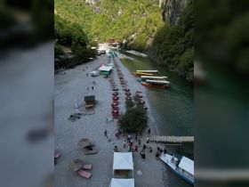 A vibrant scene at a riverside beach with groups of people enjoying the day, surrounded by lush mountains and colorful boats docked along the shore.