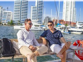 Two friends relax on the deck of a yacht, enjoying the scenic views of the city skyline and marina — a perfect setting for a leisurely day on the water.
