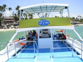 Guests gather on the deck of the Eze & Ema Crazy Boat, enjoying the vibrant atmosphere with a beautiful beach backdrop — perfect for a fun day on the water.