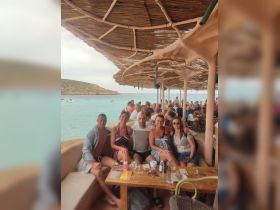 A cheerful group enjoys a meal at a beachside restaurant, surrounded by stunning turquoise waters — perfect for social gatherings and celebrations.
