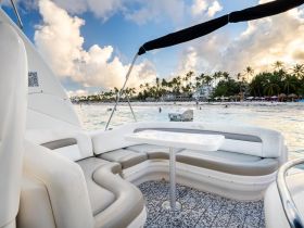 A cozy seating area inside a boat, featuring comfortable cushions and a table, perfect for relaxing while enjoying views of the beach and palm trees.