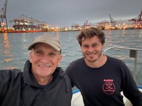 Two men smile for a selfie aboard a boat with a bustling harbor in the background, capturing a memorable moment on the water.