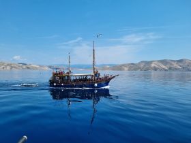 A traditional boat sails smoothly across calm waters, showcasing its elegant design against a stunning backdrop of hills and clear blue skies.