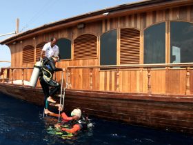 A diver ascends the ladder of a wooden boat while being assisted by a crew member, showcasing an exciting scuba diving experience in clear waters.