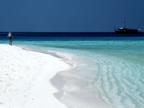 A fisherman casts his line on a pristine beach with turquoise waters and a boat anchored in the distance, capturing the essence of a serene fishing experience.