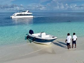 Two individuals stand on the beach near a small boat, with a larger vessel anchored in the background, creating a picturesque scene perfect for beach outings and water adventures.