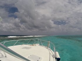 A stunning view from the bow of a boat cruising over crystal-clear turquoise waters, showcasing the vibrant marine environment and dramatic cloud formations.