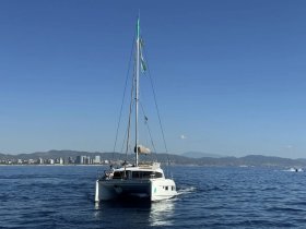 A sleek catamaran sails smoothly on the open water, showcasing its elegant design against a backdrop of coastal cityscape and mountains.