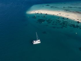 Aerial view of a catamaran anchored near a sandy beach, surrounded by clear turquoise waters — a perfect setting for a relaxing day on the water.