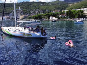 A lively scene featuring guests enjoying a sunny day on the water, with some swimming and relaxing on floating mats beside a vibrant yacht named KIGO.