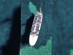 Aerial view of a sailing vessel with guests enjoying the sun on deck, while one person swims in the clear turquoise waters — a perfect scene for a day of adventure on the water.