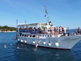 A lively group enjoys a day on the water aboard the Kuzma, with guests diving and swimming around the boat — perfect for summer parties and fun on the sea.