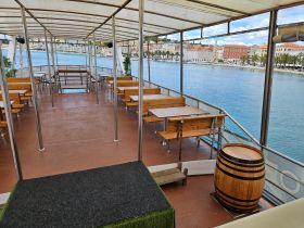 Interior view of a spacious boat cabin featuring wooden seating and tables, perfect for dining and socializing while enjoying scenic waterfront views.