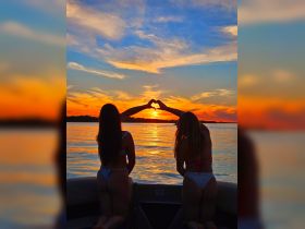 Two friends create a heart shape with their hands against a stunning sunset backdrop while enjoying a boat ride — a perfect moment for friendship and celebration on the water.