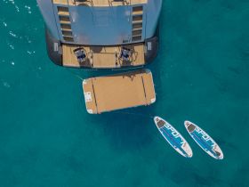 Aerial view of paddleboards and a floating mat anchored next to a luxurious yacht in crystal-clear waters, perfect for water sports and relaxation.