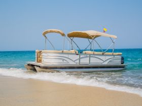 A beautiful pontoon boat rests on the sandy beach, showcasing its sleek design against the backdrop of clear blue waters — perfect for a day of relaxation and fun on the water.
